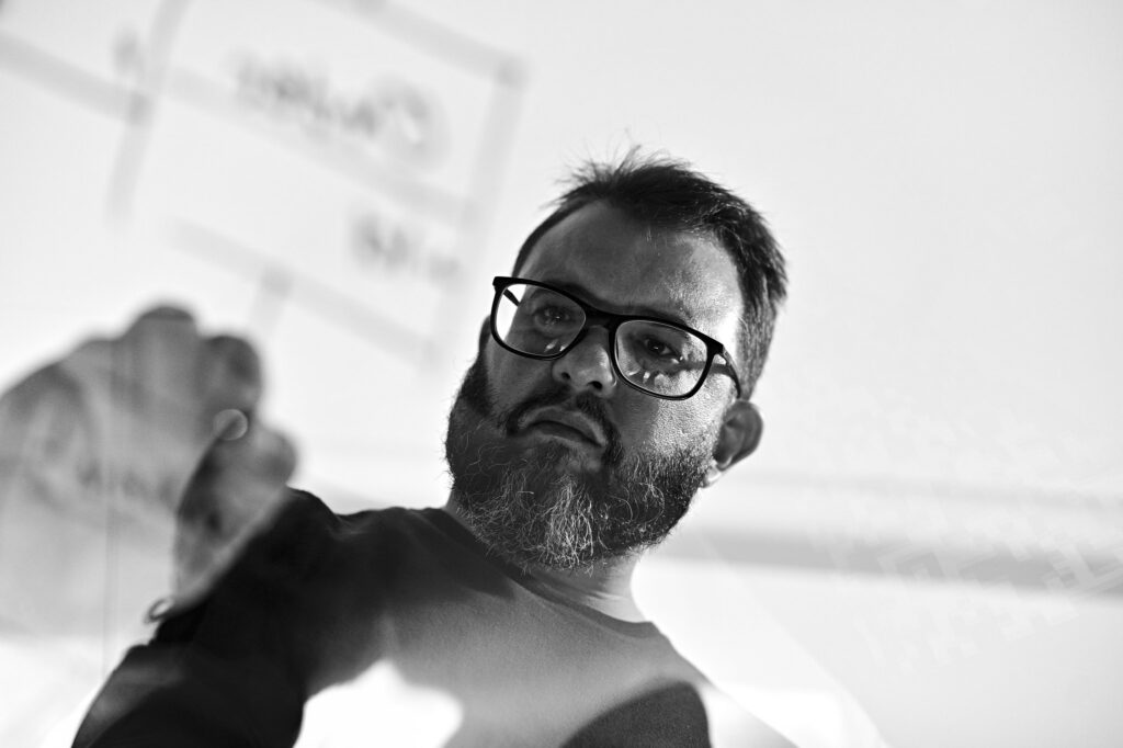 A monochrome frame of Rodolfo Mendes with the glass diagram soft above his shoulder.